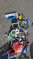 Top-down wide view of all tools and hardware items scattered on pavement showing various hand tools, screws container, sanding sheets, spray cans, tiles spacers, measuring tape, saw, and other assorted items.