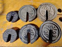 Six vintage round cast iron scale weights with semi-circular slots laid on a wooden surface. Various sizes with visible numeric weight markings.