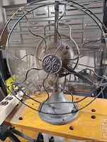 Front view showing GE logo, 4-blade fan inside protective wire cage on cast metal base