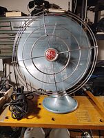 Front view of vintage General Electric fan showing blue blades behind protective wire cage and red GE logo in center hub with attached power cord.