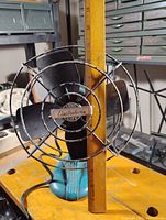 Front view of the vintage Electrohome fan showing brand badge and black blades with blue base on a workbench.