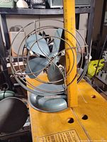 Front view of vintage blue desk fan showing blades and protective metal cage, with measuring ruler for size reference.