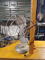 Side angle view of vintage metal fan with a ruler for scale, showing three metal blades inside a wire protective cage, sitting on workbench.