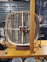 Front view of the vintage fan with a ruler to indicate size. Fan has a beige grid-style grille and a brown rectangular base.