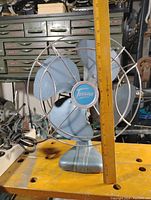 Front view of the vintage Torcan fan showing blue-gray blades behind a metal protective cage and the central Torcan logo.