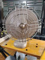 Front view showing metal fan cage enclosing three metal blades, bronze/gold finish, sitting on metal base with cord.