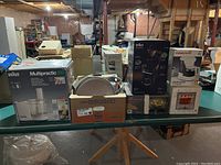 View of several boxed kitchen appliances and accessories including Braun food processor, coffee makers, and plates on a green table in a basement area.