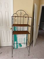 Full view of the metal and wicker bakers rack, showing its open frame, semi-circular decorative top, and three wicker shelves.