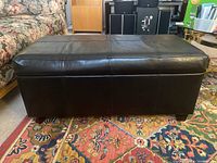 Front view of black vinyl storage bench/ottoman showing cushioned top with stitched detailing and short legs, placed on a patterned rug beside a floral couch.