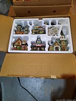 Set of six ceramic Christmas village buildings and Christmas light cords and bulbs in original packaging within a cardboard box.