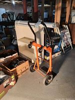 Orange hand truck/dolly with black handles and large wheels, pictured in a basement next to boxes and miscellaneous items.