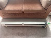 Full view of white Holmes electric baseboard heater placed on carpet in front of sofa, with power cord wrapped on one side.