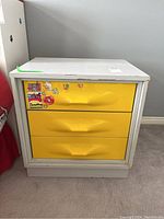 Front view of grey MDF end table with three yellow drawers and stickers.