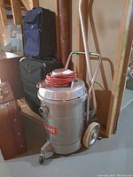 Photo of Clarke floor machine model 612 showing metal cylindrical body, handle, and power cord wrapped on top with luggage cases in background.