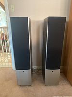 Front view of both speakers with fabric grilles on, showing silver lower baffle panels and black grilles