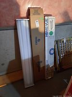 Three fluorescent light fixtures leaning against wall, two unboxed and one boxed. Boxes labeled Sylvania and Home-Vue.