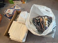 Box and bag of assorted folded fabric and plastic tablecloths, draperies, and window coverings laid on carpet.