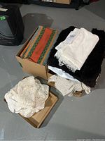Two boxes filled with various folded and piled fabrics, including a colorful striped textile, white and cream crocheted lace pieces, and off-white folded cloths.