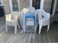 Stack of white plastic patio chairs in two different styles beside a stack of blue plastic end tables covered in plastic wrap on a wooden deck