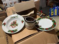 Photo of two plates and one mug from the lot, showing holiday-themed decorative motifs with red berries, pine cones, green foliage, and ribbons.