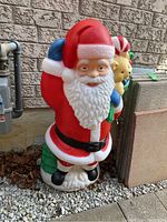 Full view of the plastic Santa Claus figure standing against a brick wall outdoors, showing the entire figure including the Santa suit, beard, and the items he carries.