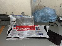 Two packs of Timberline HD roofing shingles, packaging opened, stacked on floor in basement next to some other debris.