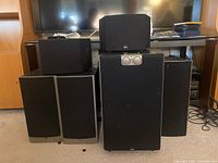 Five Athena surround sound system speakers and a large subwoofer arranged in front of a TV stand, showing the overall size and design.