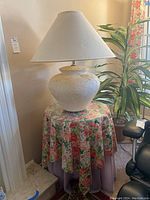 Front view of a large white textured urn-style ceramic table lamp on a round side table with floral tablecloth