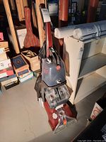 Full view of red Hoover SpinScrub 50 upright vacuum cleaner showing main unit and attached cleaning liquid tanks. Positioned on concrete floor with household items in background.