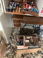 Singer sewing machine mounted in heavy oak wood table with pedal stand visible. Below table is a box containing sewing books and supplies.