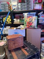Black metal shelving unit filled with cardboard boxes, bags of yarn, plastic containers, and other items. Wooden box and black metal steamer trunk also visible stacked in front.