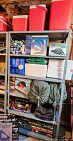Metal shelving unit with camping gear items arranged on shelves including red coolers, white Styrofoam coolers, green tarps, tent in box, sleeping bags, air conditioner, air pump