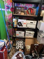 Black metal shelf with multiple cardboard boxes, craft books, some framed pictures and miscellaneous items on a floral wall background.