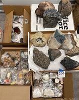 Top view of multiple cardboard boxes containing assorted rock and mineral specimens including larger geodes, agate sections and smaller bagged samples