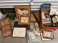 Wide view showing all items including framed sandstone art pieces, jade framed artwork, vintage axe, assorted containers, vintage ornaments, and books in boxes and on floor.