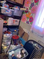 Corner storage with boxes, bins, packing materials, and yarn bags stacked near floral wallpaper and a window with sheer curtains