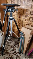 Wide view of large tripod with wooden turntable and smaller folded tripod beside cardboard box.