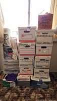 Stack of labeled and unlabeled boxes containing various sewing and fabric supplies including a container labeled 'Jean Fabric Solution Sks' and others labeled 'Office Paper' and 'Needle Work'