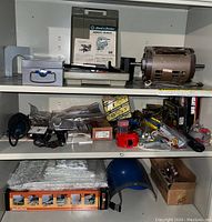 View of three shelves containing assorted handyman tools including power tool motor, staple tools, wrapped items, cables, and a blue face shield.
