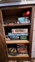 Shelves in cabinet with board game boxes visible: Spare-Time Bowling, Trouble, Fishing Game, Chopper Strike, Aurora AFX Ultra 5 Racing, Tiddly Winks, Dragon Masters, and cork dart board