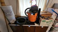 Photo showing various yard and garden tools including a Rigid shop vac with hoses, rolled insect screening, cardboard boxes of paper bags, white 5-gallon buckets, and Scott's fertilizer spreader