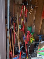 View of various yard tools including shovels, rakes, clippers, brooms, and bags of potting soil. Part of a bike frame visible in the corner.