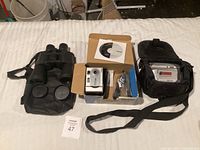 Lot items laid out on white surface: binoculars in case, boxed digital camera with software CD, another camera in carrying case.