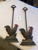Pair of cast iron rooster andirons with tall rods and decorative handles placed on white cloth backdrop.