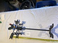 Full length photo of cast iron weathervane showing rooster figure, base and measuring tape indicating about 30 inches long.