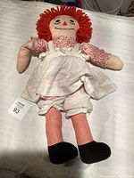 Front view of the cloth doll with red yarn hair, floral blouse, white pinafore, bloomers, striped legs, and black shoes placed on a white surface.