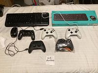 Multiple gaming controllers and keyboards displayed on a white surface. Controllers include Xbox wireless controllers, a wired PlayStation-style controller, and mice. Keyboards include Microsoft and Logitech models, one Logitech keyboard in original box.