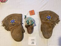 Top view of two pairs of brown leather moccasins with decorative beadwork and fringes; miniature moccasin container with small figurine in center