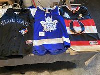 Three sports jerseys laid side by side including Toronto Blue Jays, Toronto Maple Leafs, and Ottawa Senators.