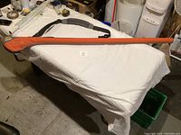 Orange and gray Plano 65-inch fly rod holder case lying on a table, showing full length and shape with shoulder strap.